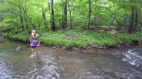 Bikini clad mature woman hiking through natural mountain stream or creek in forest, captured with steadicam