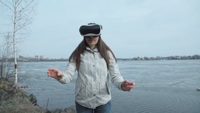 Attractive young woman in warm white jacket uses virtual reality glasses while standing next to a pond covered with a thin crust of ice in spring day.
