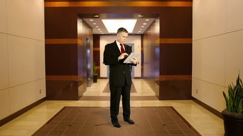 Mature businessman standing in middle of office building lobby area working on electronic tablet.