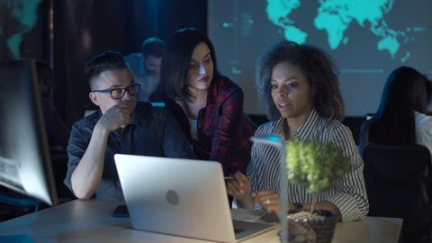 Mixed racial people working in modern office with low light interior, two women and man in front of computers discussing something