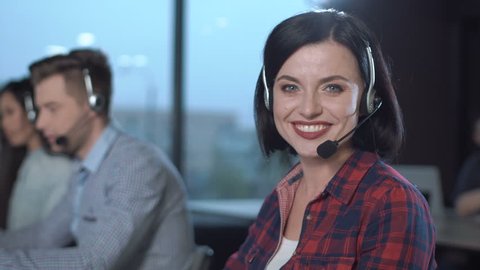 Call center operators at work, with young woman in earphones and mic headset in foreground talking to client. Close-up side portrait, copy sace