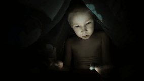 Child with a tablet on the bed, under the blanket