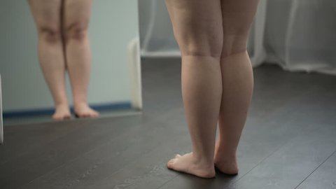 Legs of excess weight woman with flat feet and cellulite, obesity problems. Fat female turning around in front of mirror, looking at her body, insecurities, examining stretch marks, dieting. Plus-size