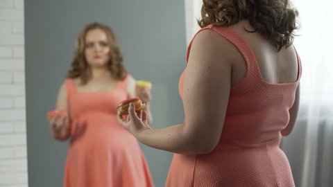Depressed overweight lady chewing donuts in front of mirror, eating disorder