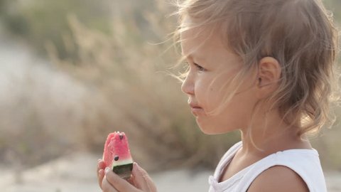 Close-up portrait of small cute baby girl eat red watermelon at sunrise, outdoor