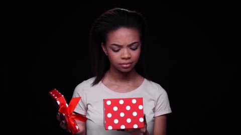 Excited young afro american woman opening gift box and looking disappointed isolated on a black background