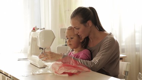 Young attractive mother working on sewing machine with her little cute daughter. Mother teaching daughter new knowledge.
