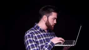 Young surprised bearded man working on laptop while standing isolated over black 