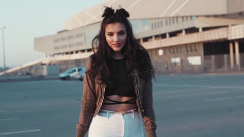 Attractive young woman in a stylish outfit turns to camera and smiles seductively, continues walking away from the camera. Fashionable look, tight white trousers, bomber. Fashion blogger.