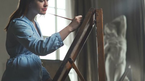 A young woman paints a painting on a canvas, standing behind an easel, in an atmospheric workshop with panoramic windows and an antique statue. Art academy or drawing school. Inspiration. Talent.