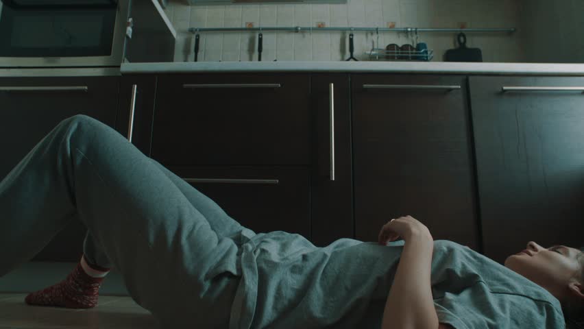 Young caucasian fast reflections woman in grey clothes lying on kithen floor sits up and catch falling red glass, slow motion