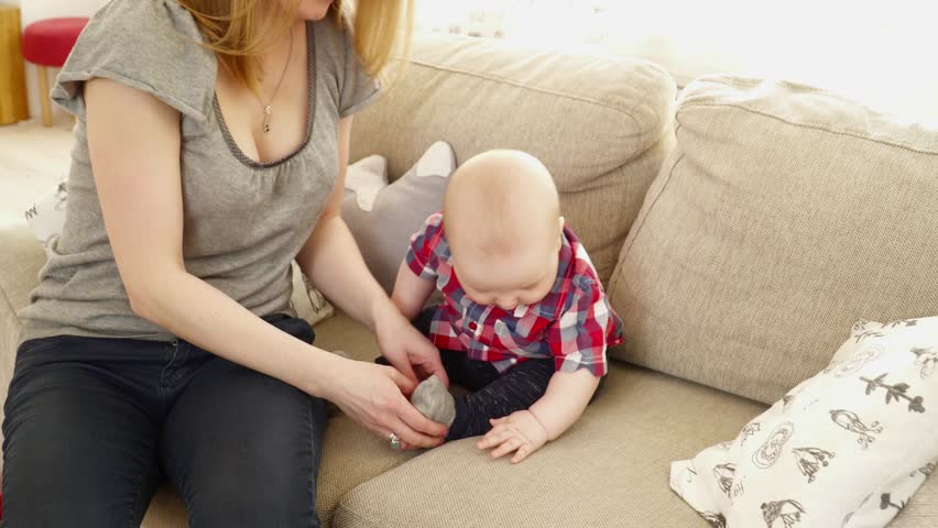 Young mother puts the socks on to her 6 months baby boy on sofa