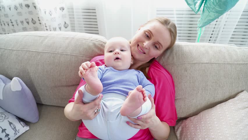 Pretty mother playing with 6 months son's legs laying on the sofa