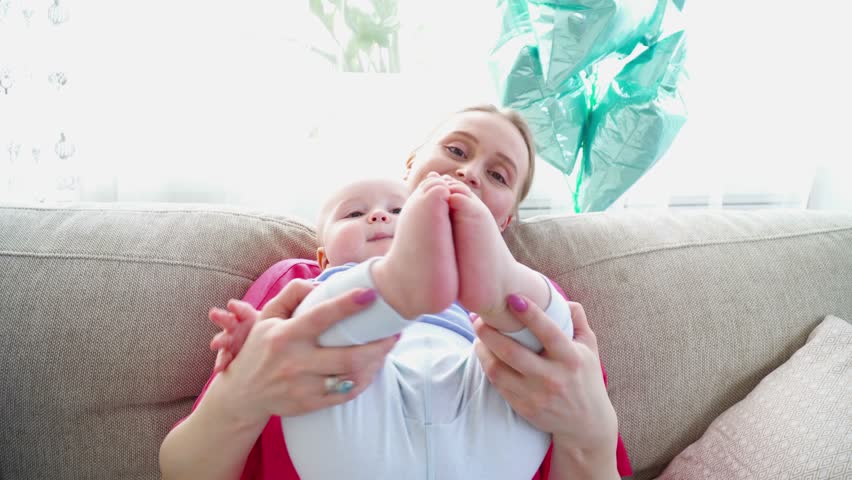 Young mother playing with 6 months son's legs laying on the sofa