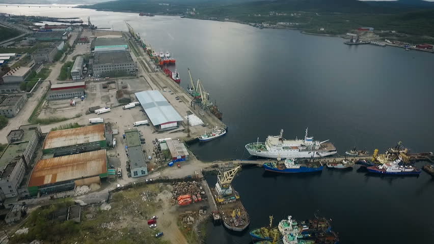 Sea Port Top View. Different Size Boats Staying in Bay. Heavy Crane Moving. Storage Building on Shore. Ships With Load at Moorage. Wide Shot