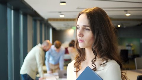portrait of positive attractive brunet young working woman who look and smile to camera holding blue folder with business papers staying on front of working people in light office with big windows