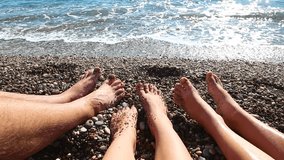 Closeup of barefoot people sitting on beach near sea water. Family of mom, dad and son enjoy their vacations on seaside in summer. Point of view real time full hd video footage.