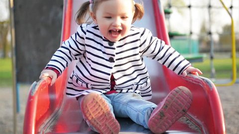 Funny Cute Girl with Two Ponytails is Playing on Red Slide. Joyous Female Child in Striped Jacket Having Fun on Playground in the Park.
