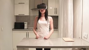 Beautiful brunette woman in white top wearing in VR glasses wiping utensils, plates and cups, putting plates on shelf. Young girl cleaning at kitchen, playing game in cyberspace.