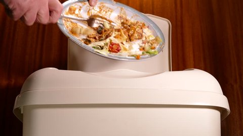 A man’s hand scraping uneaten food waste from a plate into a plastic trash can/bin.