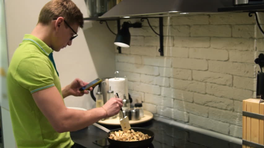 Young man cooking dinner and browsing social media on smart phone. He is always online