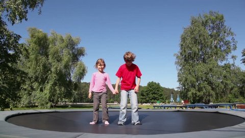 Children boy with little girl hold hands and jump on trampoline in park at summer day