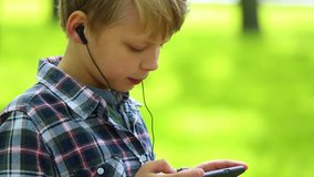 Closeup portrait of blond cute boy of 10 year old standing in spring or summer park and listening to music happily. Kid uses black headphones and modern smartphone. Real time full hd video footage.
