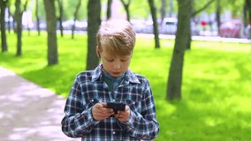 Portrait of blond cute boy of 10 year old standing in spring or summer urban park and listening to music happily. Kid uses black headphones and modern smartphone. Real time full hd video footage.