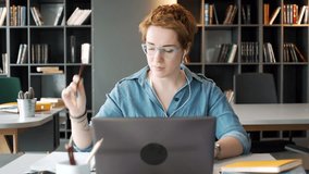 young working red haired manager take part in online video conference call using laptop computer, making notes in notebook concentratedly, sitting in office space during day