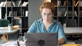 young working red haired manager take part in online video conference call using laptop computer, making notes in notebook concentrated, sitting in office space during day