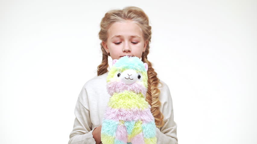 Cute beautiful Caucasian girl with luxurious long blonde hair standing on white background and softly cuddling fluffy lama toy in slowmotion