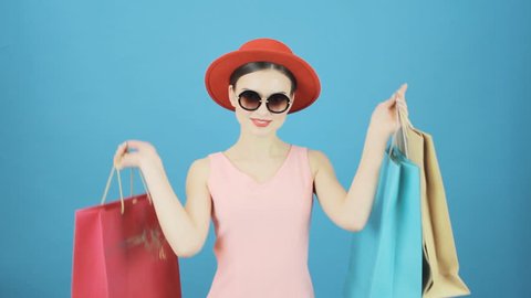 Portrait of Smiling Brunette with Red Hat and Sunglasses on Blue Background in Studio. Happy Woman Holding a Lot of Shopping Colorful Bags. Seasonal Sale Concept.