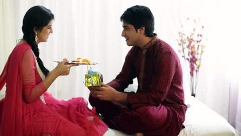Zoom in shot of a teenage girl holding a puja thali and her brother sitting with a present in front of her
