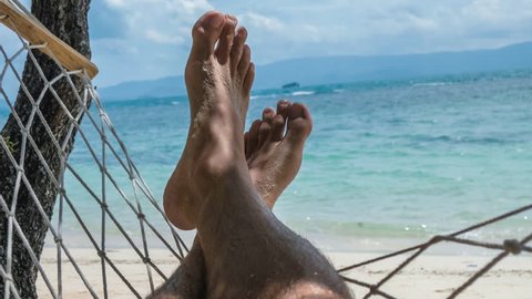 Swinging bare man feet with beach white sand swinging in a hammock on vacation in front of the blue ocean.
