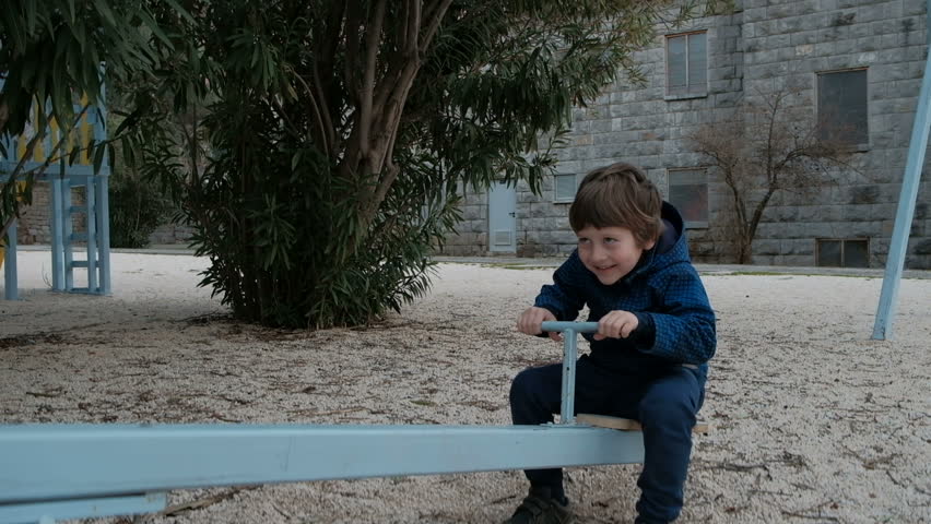 Small boy riding seesaw in playground in winter. Nobody else. Toddler with dark hair in sneakers, blue trousers, plaid coat without cap, gloves sit on wooden plank as seat in iron carousel, he move up