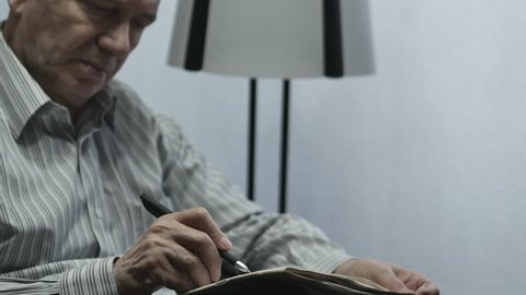 Elderly Man Reading And Writing In The Newspaper On The Couch