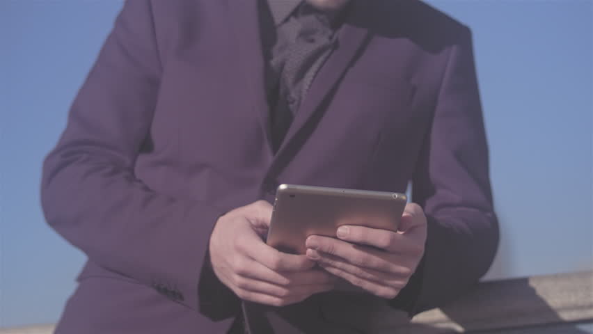 Panning shot of a young business man leaning up against a rail, while using his tablet. He smiles at the camera