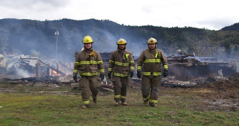 Slow-motion shot of three firemen walking towards the camera with a smoldering structure in the background