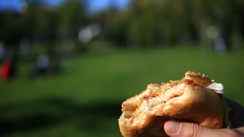 A man is eating fast food on the street. He wears a hamburger and eats it. Against the background of a blurry city street