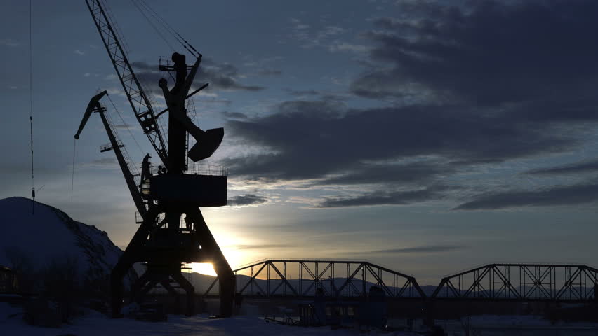 Time lapse of beautiful colorful dusk on a river in winter. Silhouettes of barge, reeds, cranes and railway bridge across the river. Reflections of lights on a water.