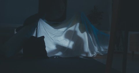 Стоковое видео: Silhouette of little girl reading a book inside a blanket fort in the evening, lit by a lamp from inside. 4K UHD RAW edited footage