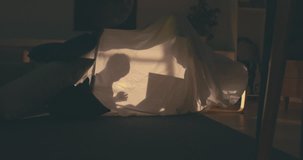 Silhouette of cute girl reading a book to her little brother inside a blanket fort in the evening, lit by a lamp from inside. 4K UHD RAW edited footage