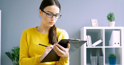 Young attractive business woman in glasses stands at the office and writing notes in notepad, thinking and looking at the camera. Portrait shot of businesswoman