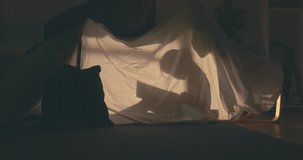 Silhouette of little girl reading a book inside a blanket fort in the evening, lit by a lamp from inside. 4K UHD RAW edited footage