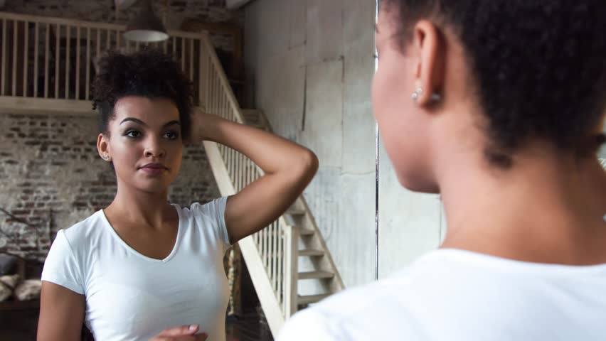 Attractive african-american girl corrects hair standing in front of a mirror.
