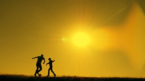 Father and son playing football in the park at sunset, silhouettes against the backdrop of a bright sun, slow-motion shooting