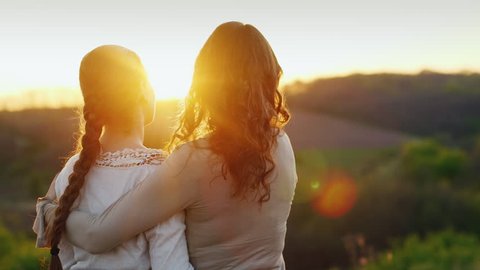 Mom gently kisses her daughter's temple. Together they admire the beautiful sunset. Back view