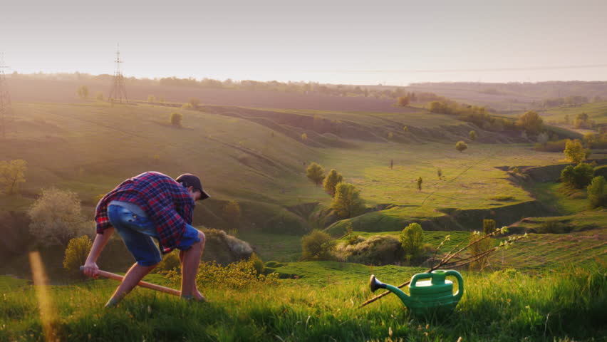 A young guy 17 years old plant a tree. Digging a hole with a shovel, next to an apple tree seedling. Against the background of a beautiful landscape . Work of volunteers, environmental protection