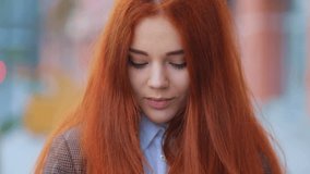 Amazing red-haired girl in business style wear coquettishly looking into the camera with slightly closed lips, urban landscape Steadicam slow motion video