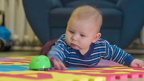 Baby With Blue Eyes and in Striped Jacket Lies on the Rug in the Room. he Plays With a Toy of Green Color. he Tries to Reach Out With His Hand to Toy.